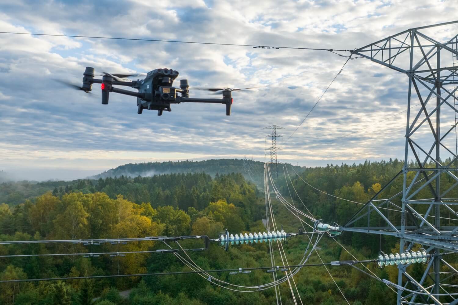 Inspection drone hovering beside a high-voltage transmission tower and insulators above forested hills